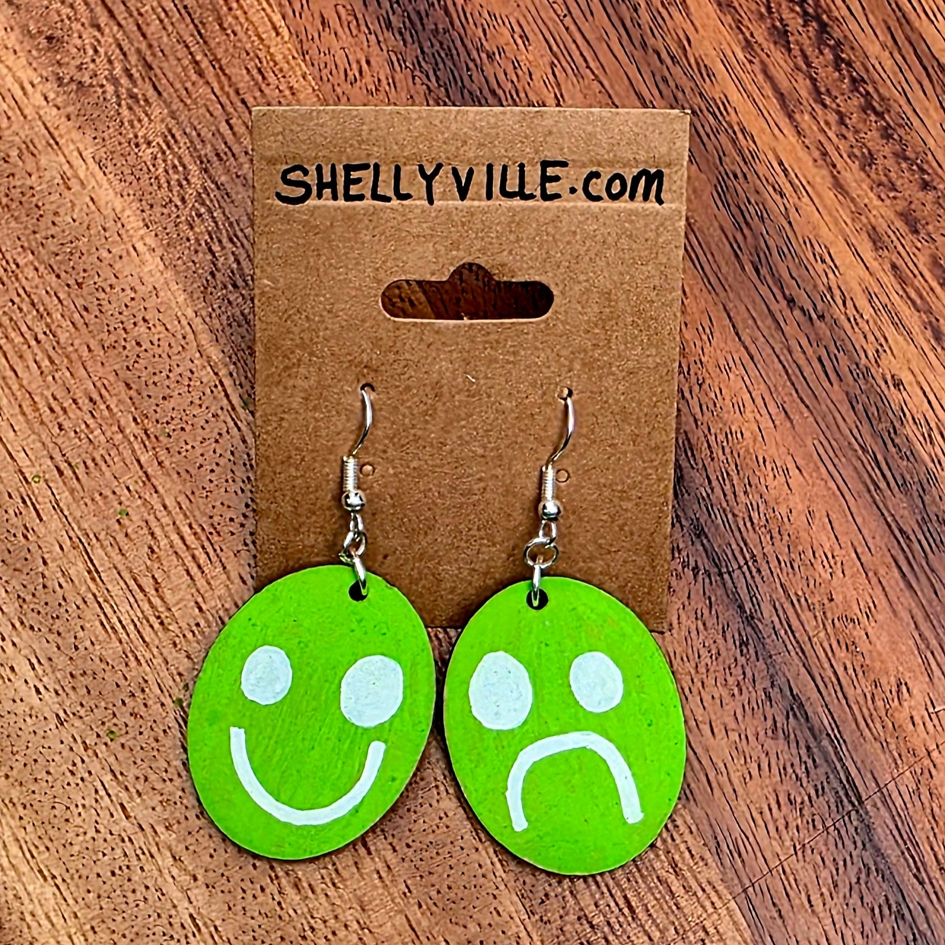 Green smiley face and frowny face earrings on a Shelly Yile branded card against a wooden background.