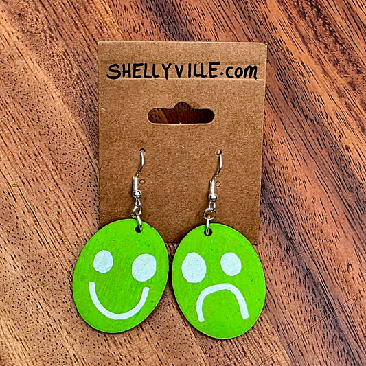 Green smiley face and frowny face earrings on a Shelly Yile branded card against a wooden background.