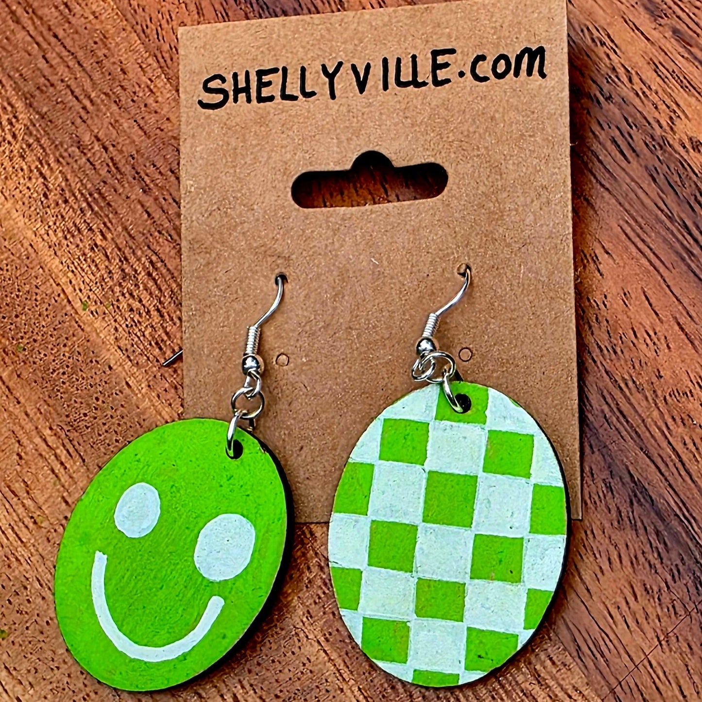 Green smiley face and checkered earrings on a Shellyville.com card against a wooden background