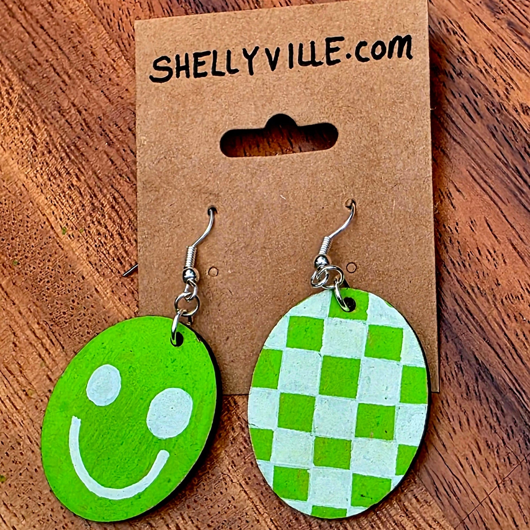Green smiley face and checkered earrings on a Shellyville.com card against a wooden background