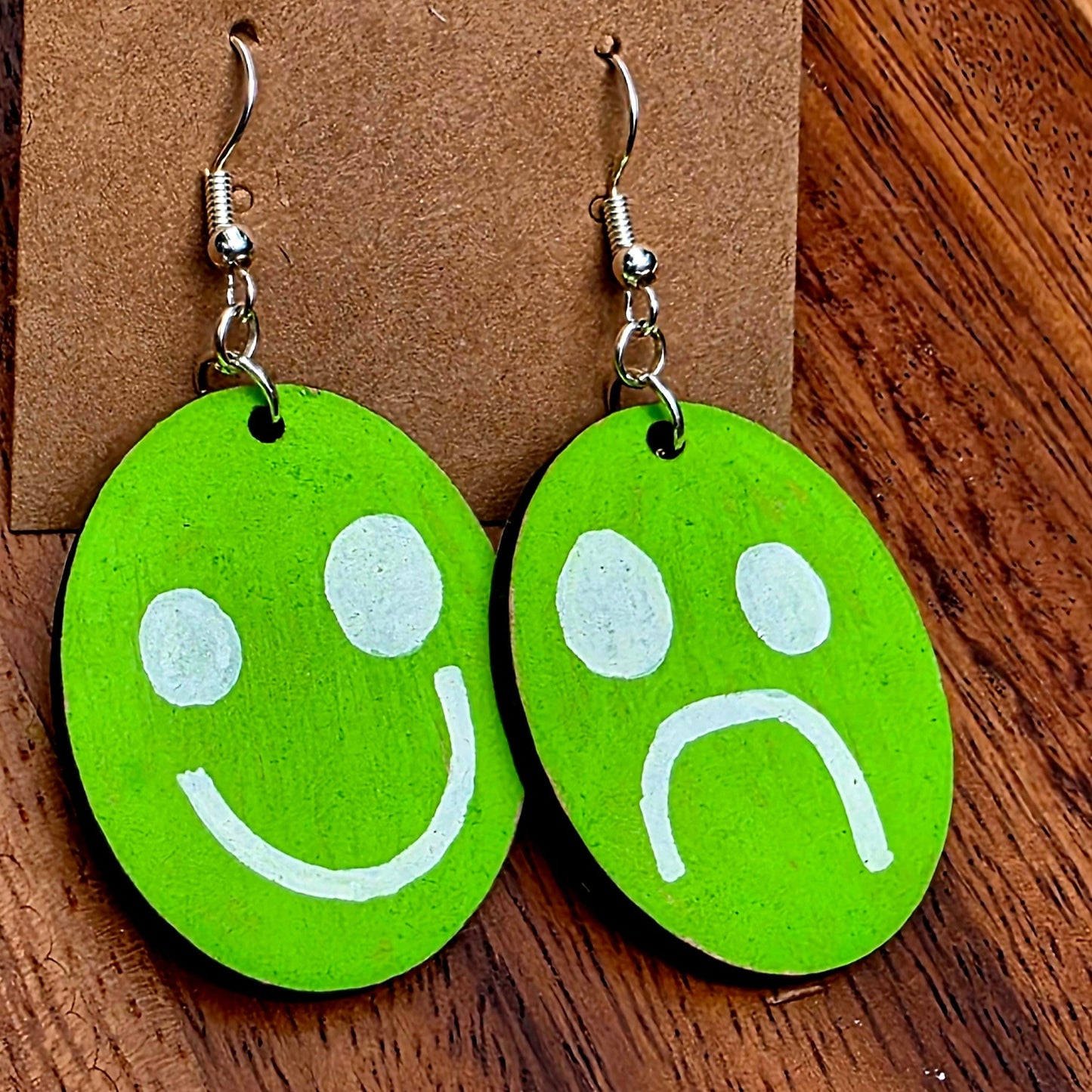 Green smiley face and sad face earrings on a Shellyville.com card against a wooden background