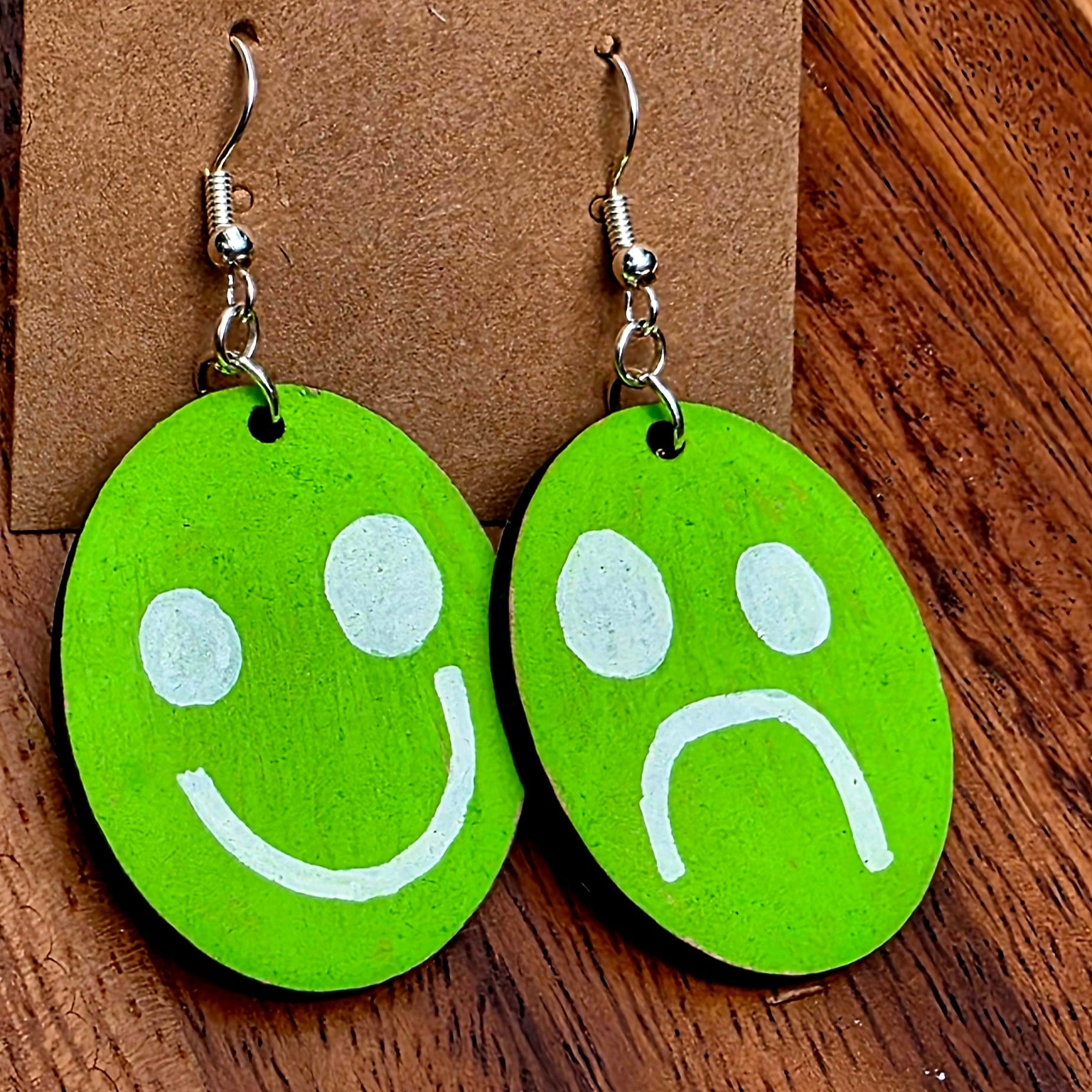 Green smiley face and sad face earrings on a Shellyville.com card against a wooden background