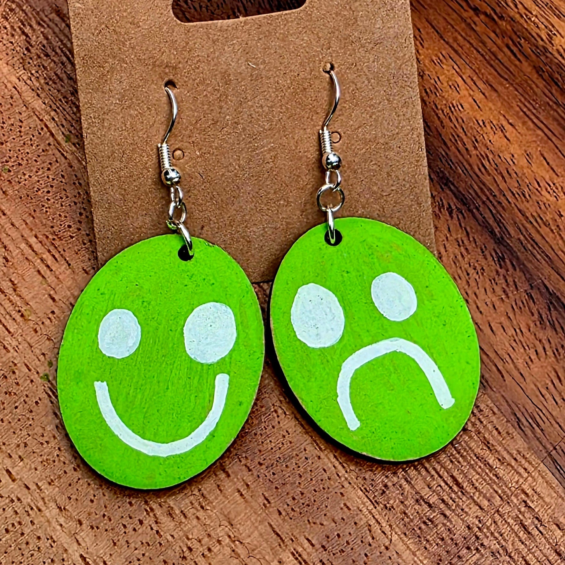 Green smiley face and sad face earrings on a Shellyville.com card against a wooden background